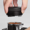 A hand holds a black precision espresso distribution tool above a metal grinder.