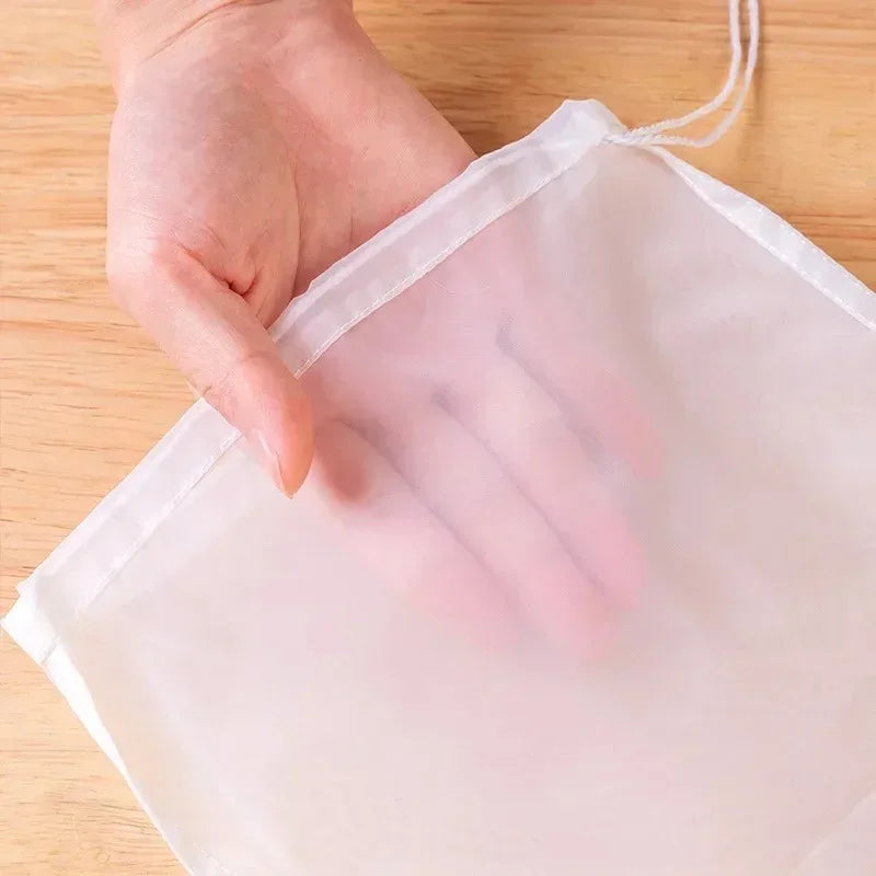 Una mano sostiene una bolsa coladora de malla reutilizable transparente hecha de tela blanca sobre una superficie de madera.