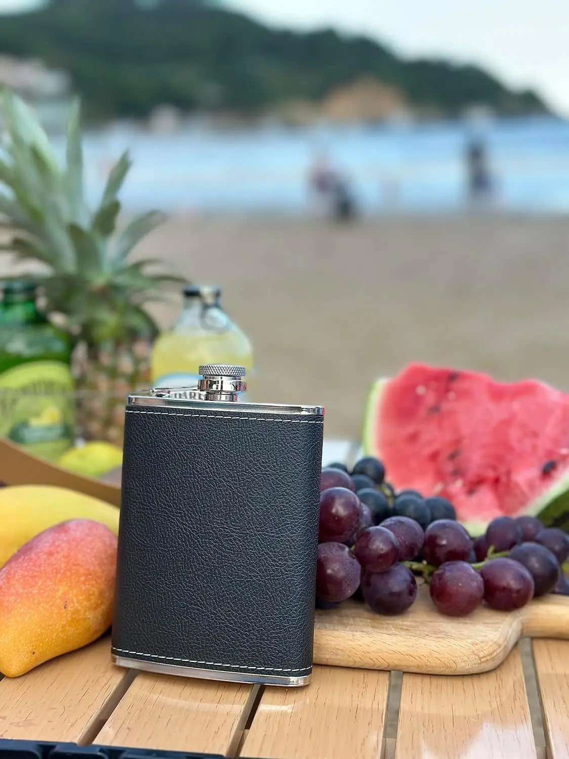 A stainless steel whisky flask with a leather cover sits on a wooden surface surrounded by fruit and a tropical background.