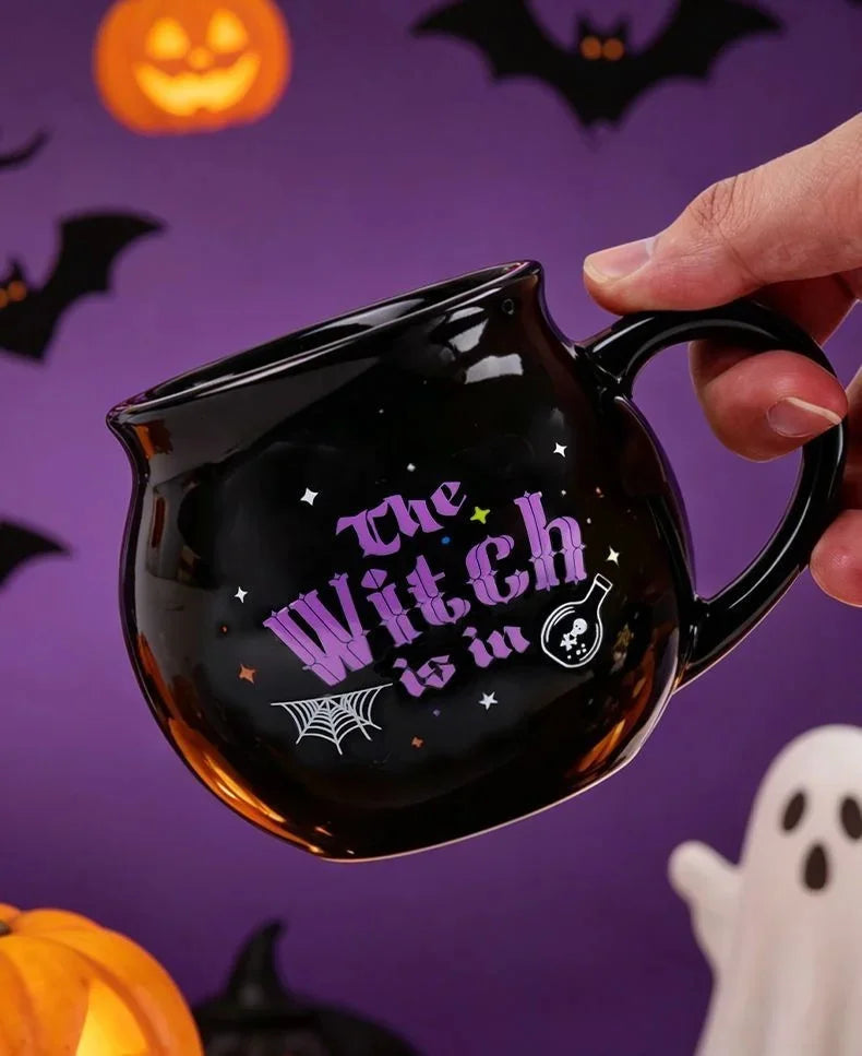 A hand holds a black cauldron mug with purple text and a witch symbol against a purple Halloween background.