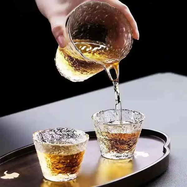 A hand pours amber tea from a glass into another small glass on a dark tray.