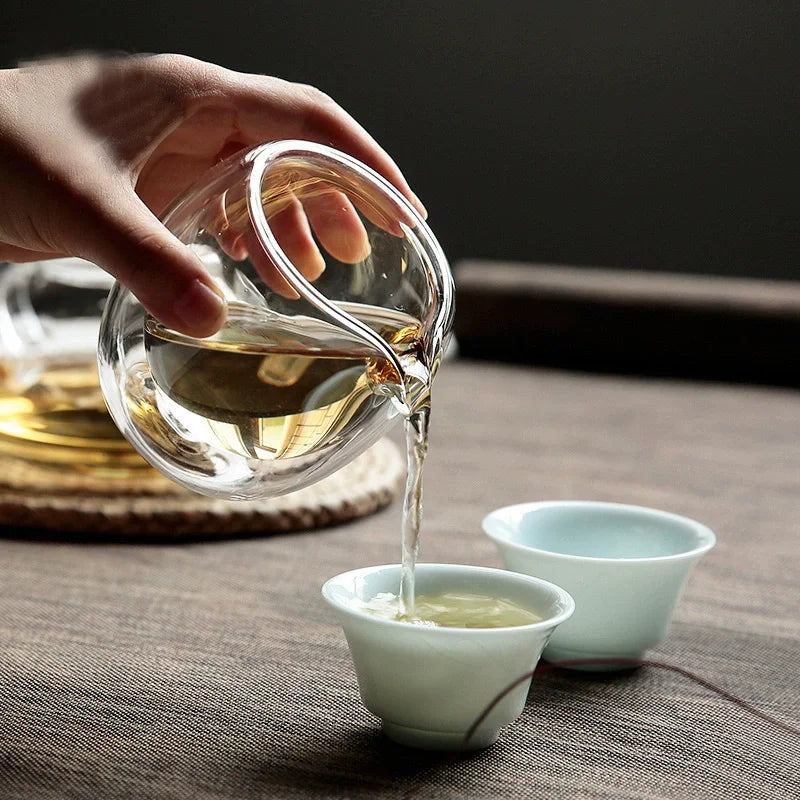 A hand pours amber tea from a glass pitcher into small white cups on a dark wooden surface.