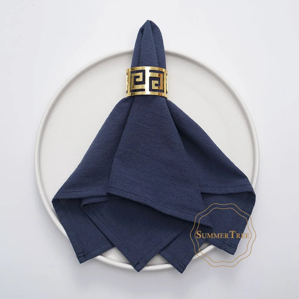 A navy blue cotton table napkin folded and tied with a gold geometric ring on a white plate.