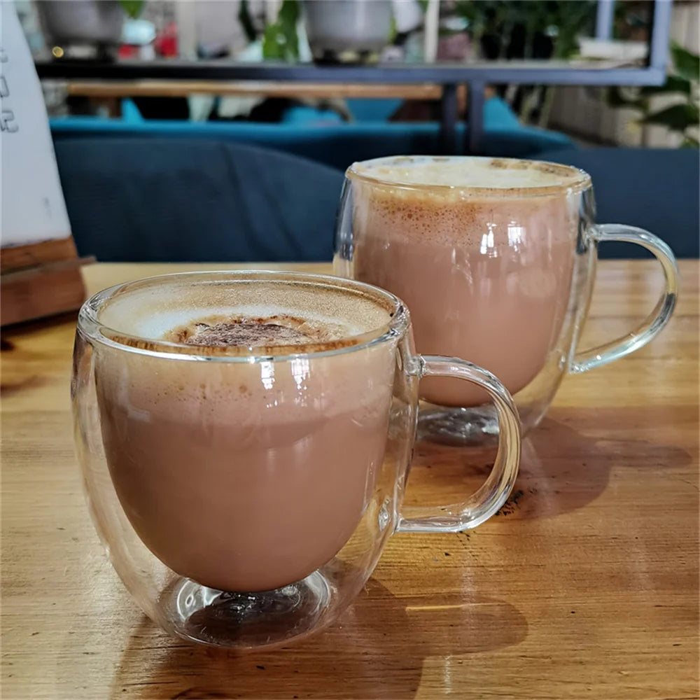 A clear double-walled glass coffee mug filled with a light brown beverage topped with foam, resting on a wooden table.