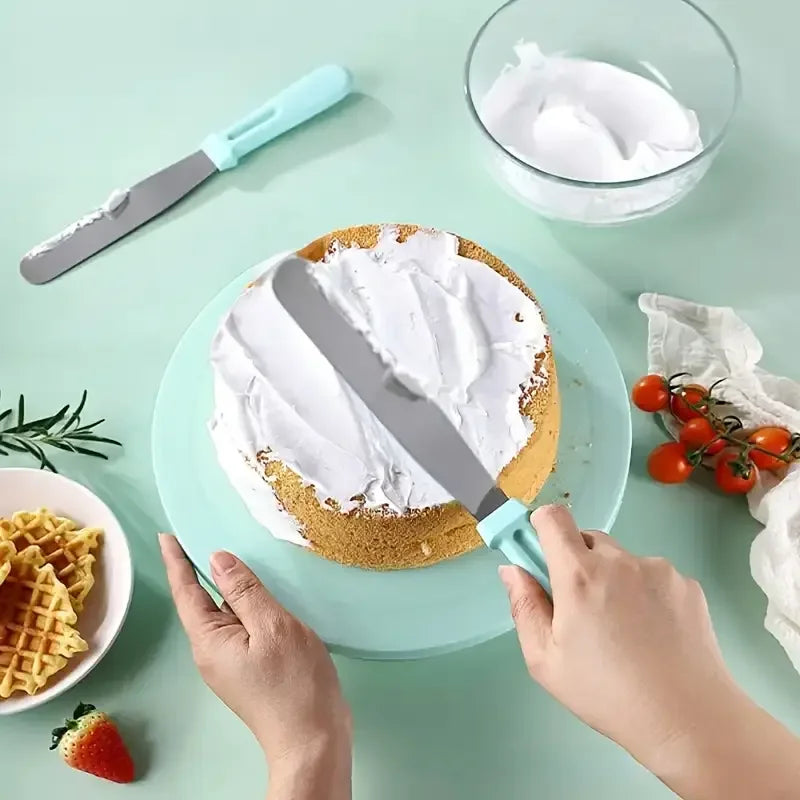 Hands are decorating a cake with white frosting using a metal knife and a turntable on a light green surface.