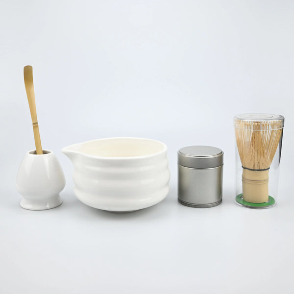A traditional matcha tea set featuring a white ceramic bowl, a bamboo whisk, and a metal container with a bamboo brush.