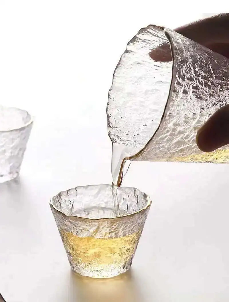 A hand pours golden liquid from a glass into another elegant glass for a tea ceremony.