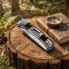 A silver vintage engraved folding utility knife rests on a wooden log next to an old book and greenery.