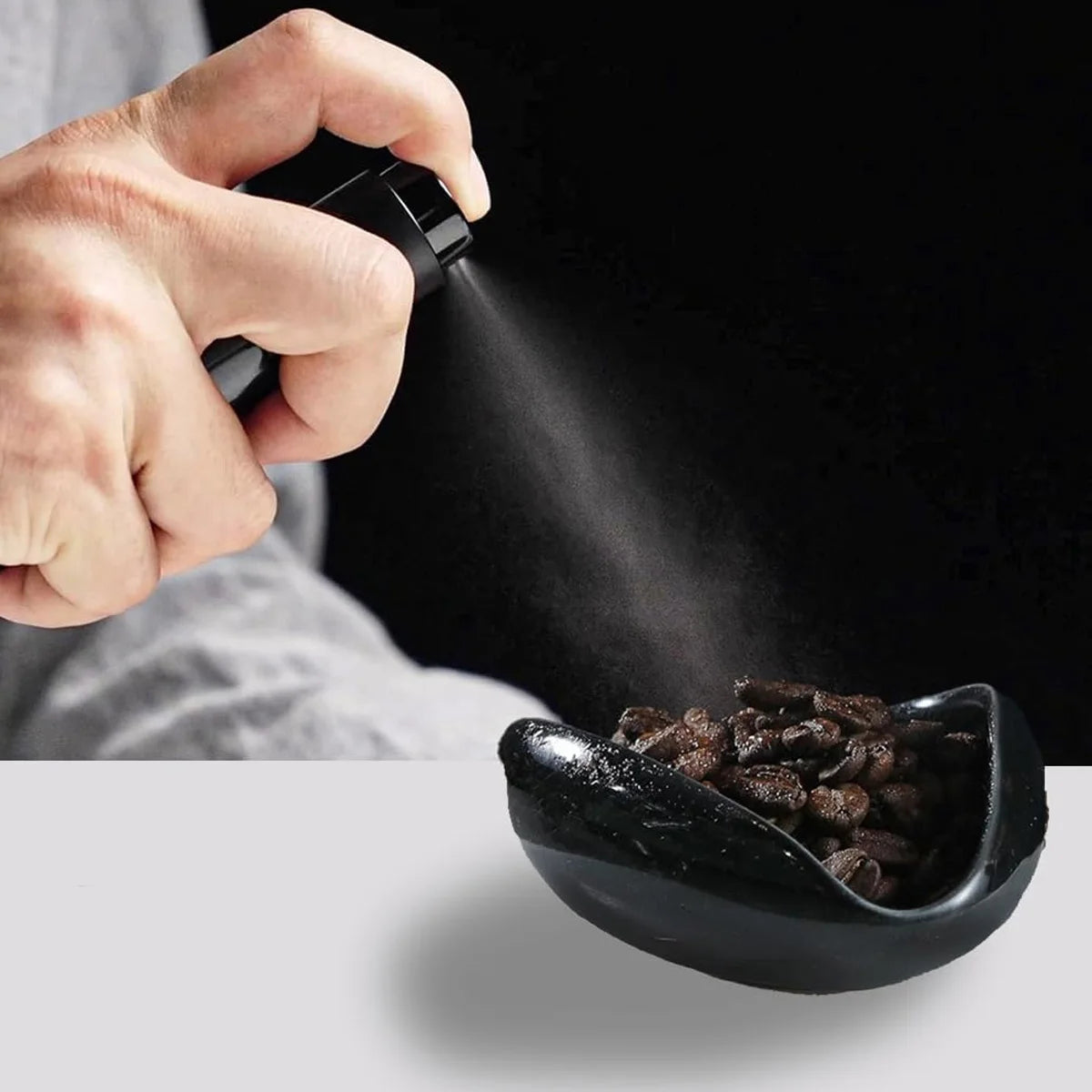 A hand is using a black ceramic coffee dosing and spray kit to dispense a fine mist onto coffee grounds.