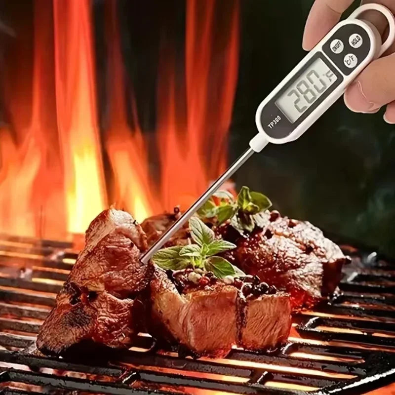 A hand holds a digital kitchen food thermometer with an instant read probe measuring meat on a grill.