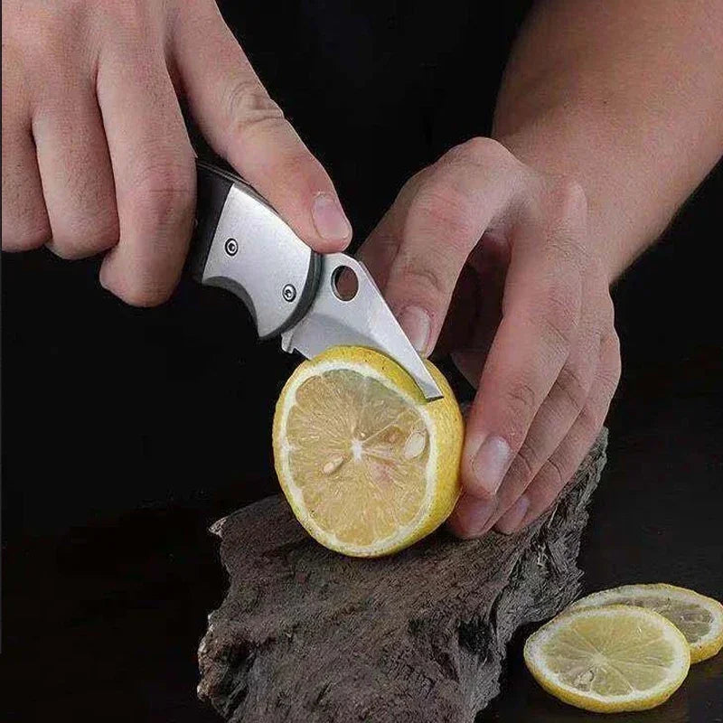 A hand is using a stainless steel mini folding utility knife to cut a slice of lemon on a piece of wood.