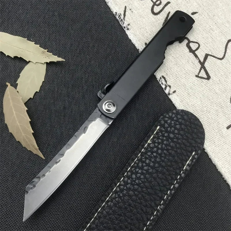 A black folding pocket knife with a silver blade is shown next to a dark fabric and a leather sheath.