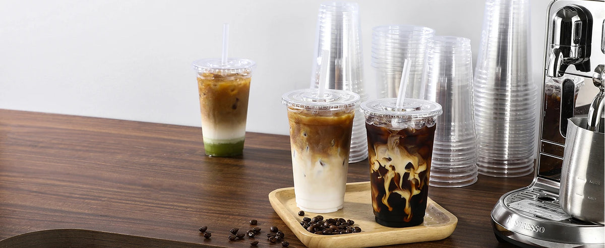 A close-up of iced coffee drinks in clear plastic cups with lids and straws sitting on a wooden table.