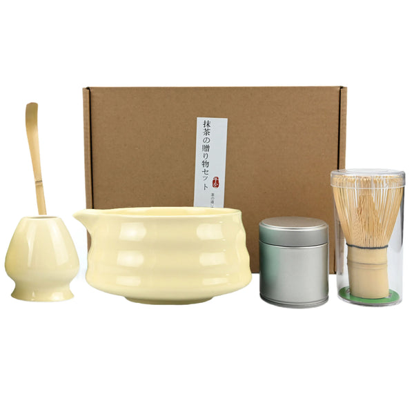 A traditional matcha tea set includes a light yellow ceramic bowl, a wooden whisk, and a bamboo tea brush in a cardboard box.