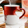 A red ceramic mug with a heart handle is shown pouring liquid, set on a white surface next to a glass of juice.
