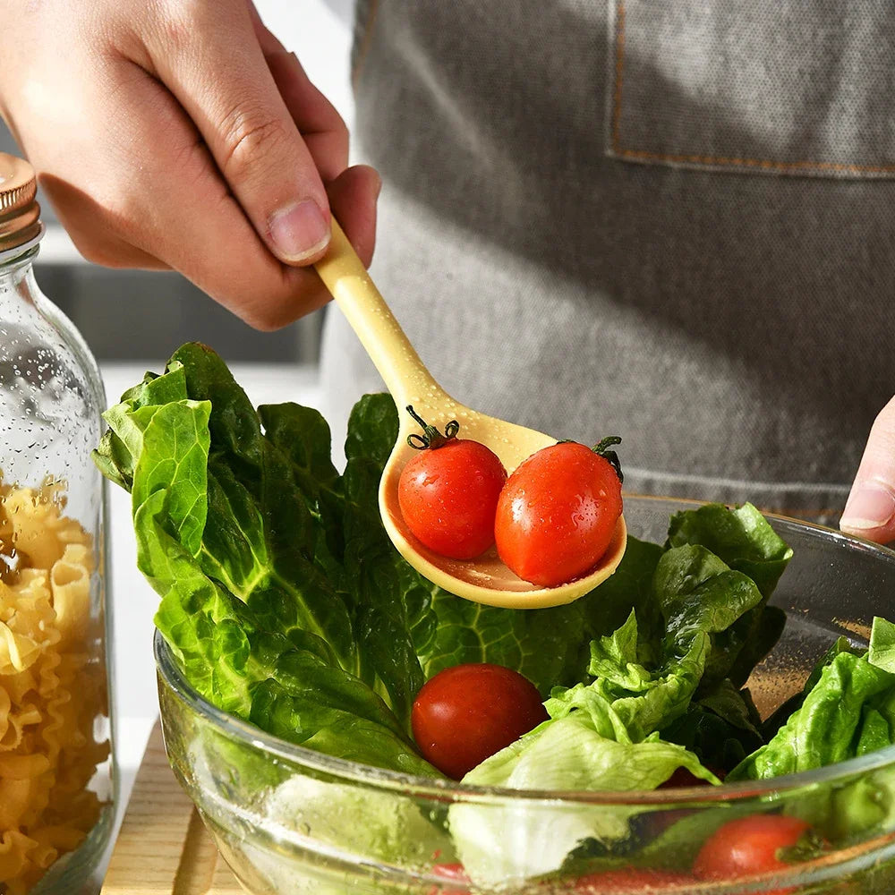 A hand uses a silicone heat resistant spoon to serve tomatoes and lettuce in a glass bowl.