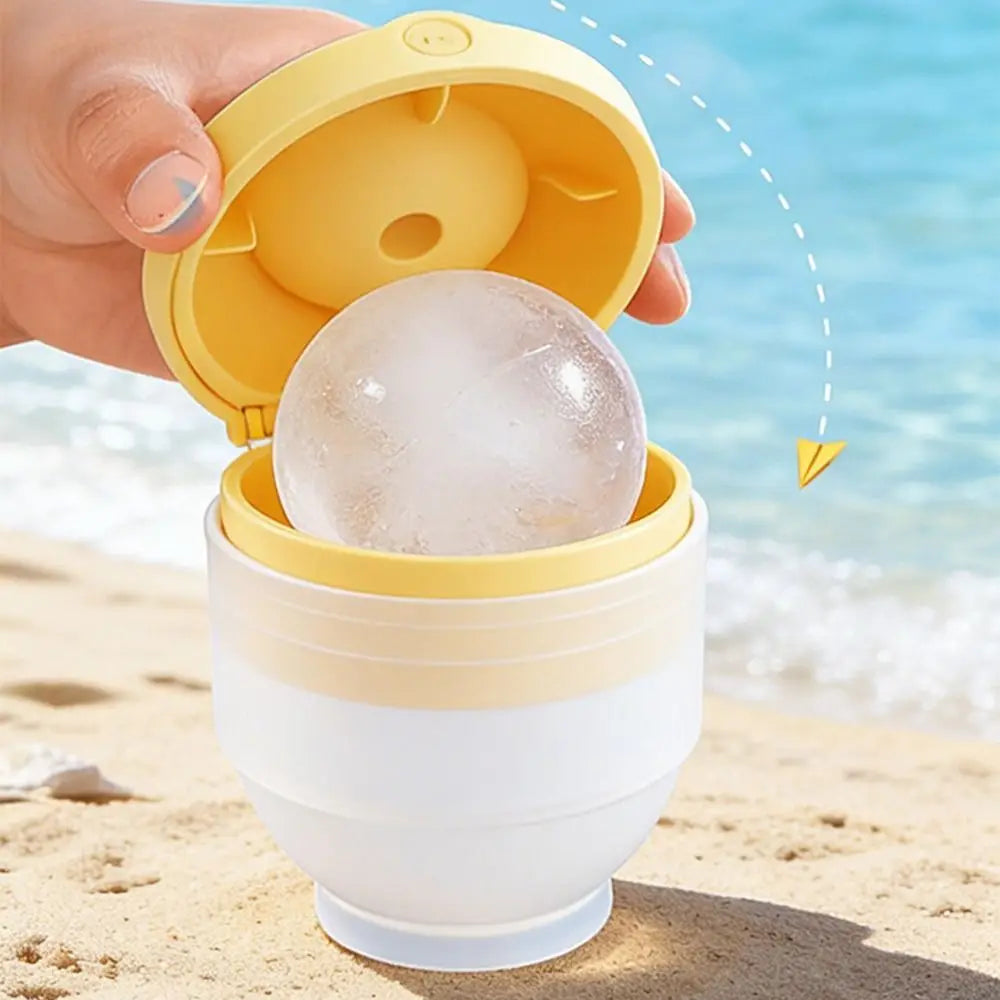 Una mano sostiene un molde de silicona amarillo abierto que contiene una bola de hielo transparente en una playa arenosa cerc