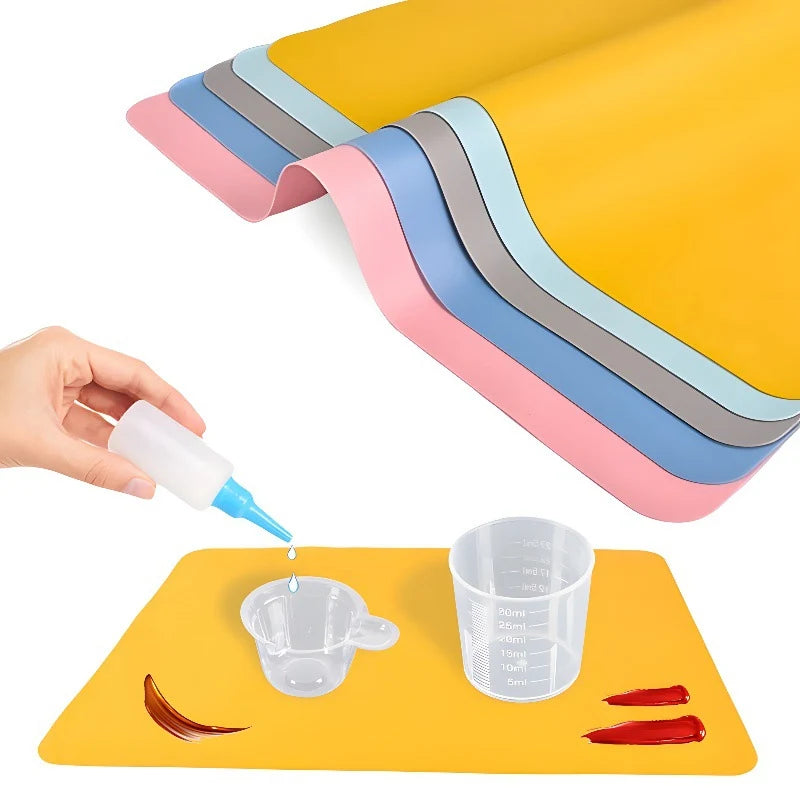 A hand is dipping a silicone mat with a dropper onto a yellow craft mat featuring layered pink and gray silicone sheets.