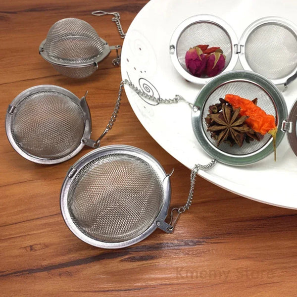 Stainless steel tea infuser balls with chains holding loose leaf tea, spices, and herbs on a white plate.