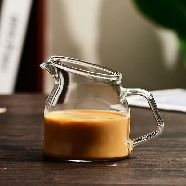 A clear glass serving cup filled with a light brown liquid, likely milk or coffee, sitting on a dark wooden surface.