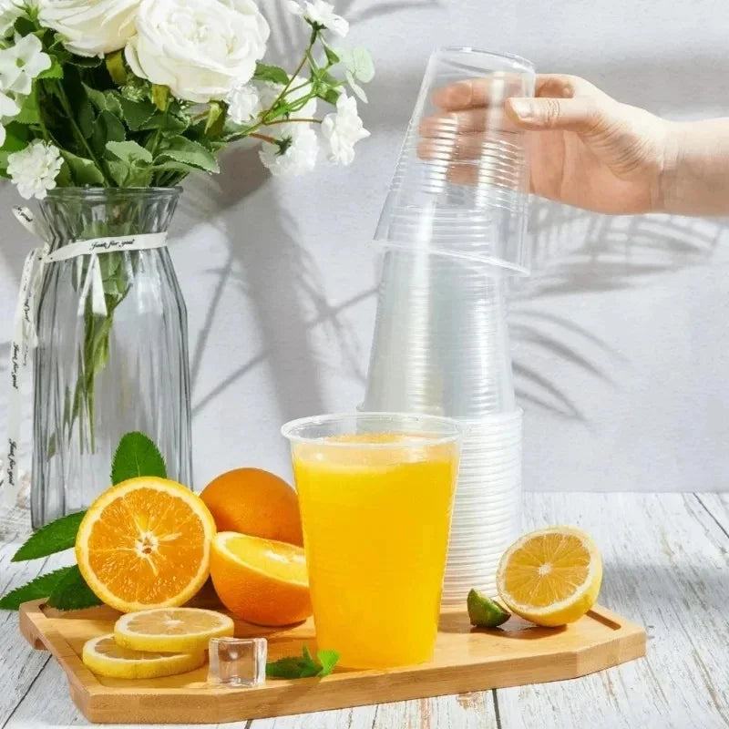 A hand pours orange juice from a clear plastic cup into a glass, with fresh citrus fruits and a glass pitcher in the backgrou