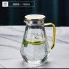 A clear glass pitcher with a gold handle and white lid, filled with green leaves.