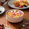 A pink bowl filled with colorful cereal and pink and white candies sits on a wooden table next to a plate of food.