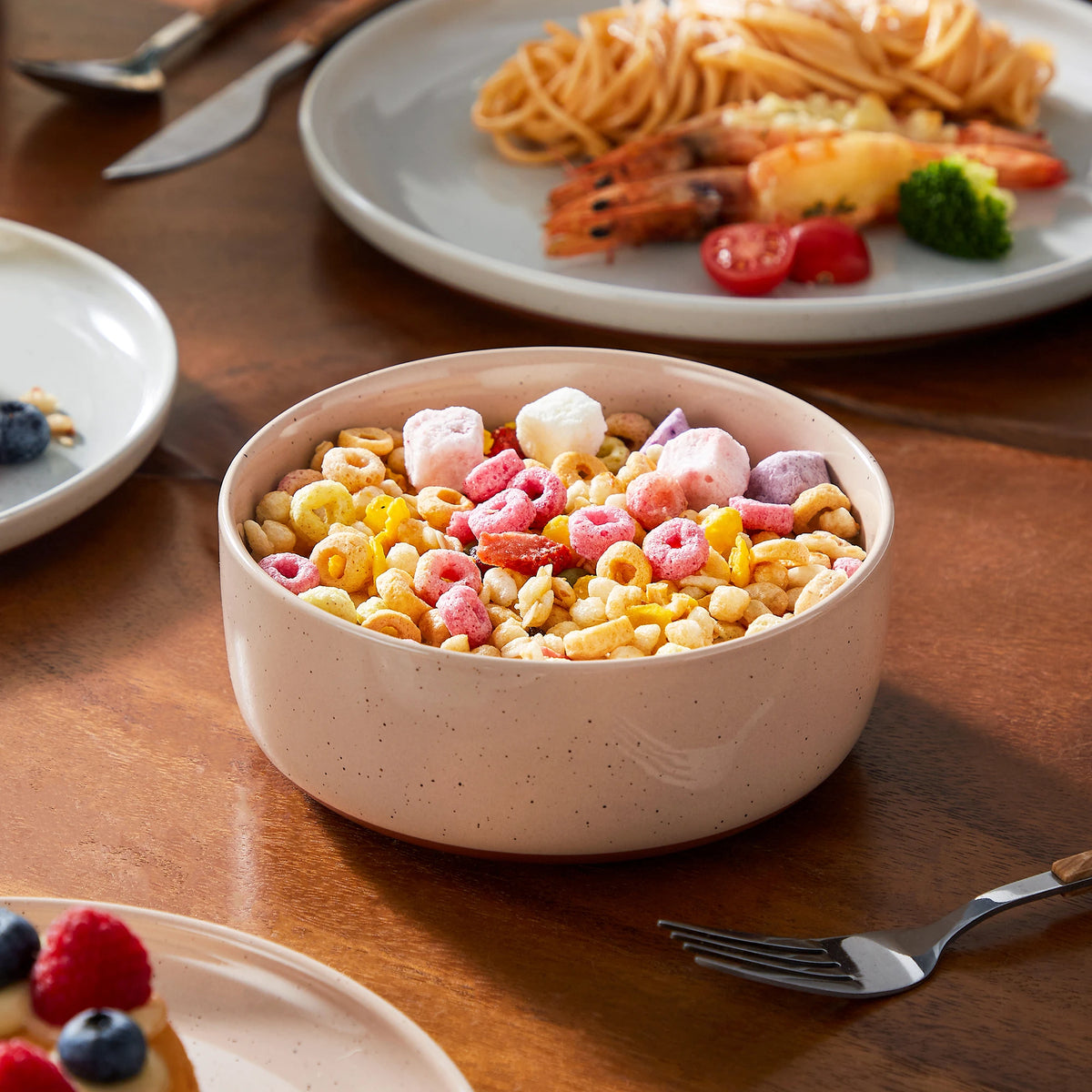 A pink bowl filled with colorful cereal and pink and white candies sits on a wooden table next to a plate of food.