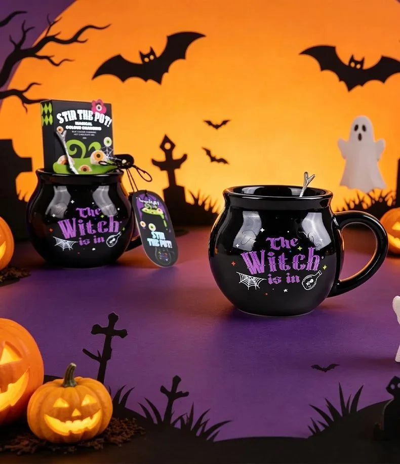 A black gothic cauldron mug with a witch design sits on a purple surface surrounded by Halloween decor.