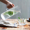 A hand pours clear liquid from a glass pitcher into a mug, showing fresh cucumber slices and mint leaves.