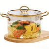 A clear glass cookware pot with a lid and double handles filled with cooked shrimp, corn, and vegetables on a wooden board.