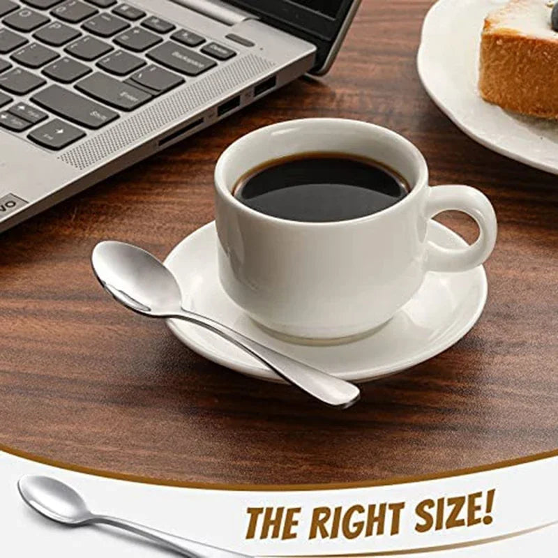 A set of stainless steel espresso spoons resting on a saucer next to a cup and a laptop.