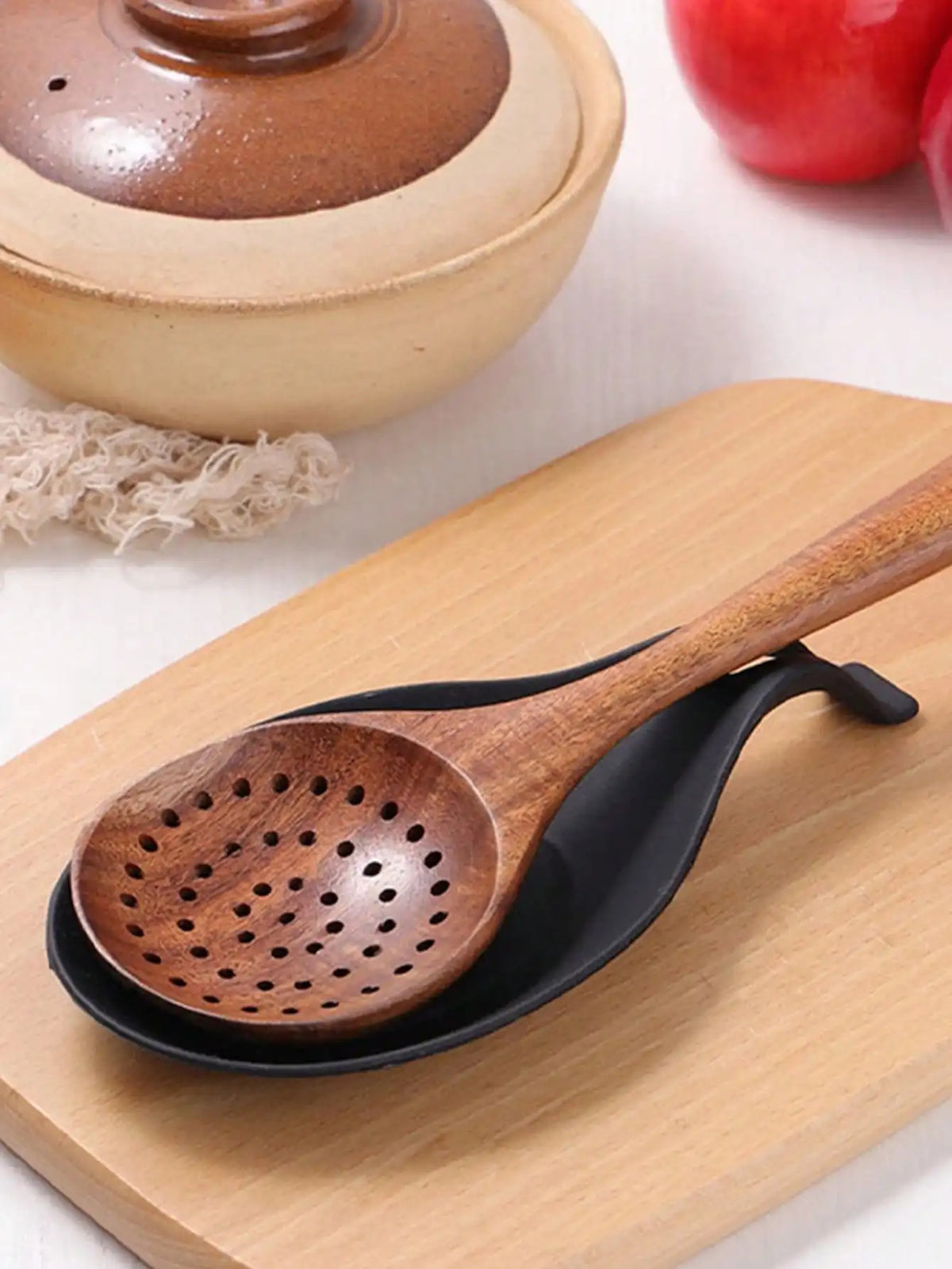 A wooden spoon rests on a black dish on a light wood cutting board next to a ceramic pot.