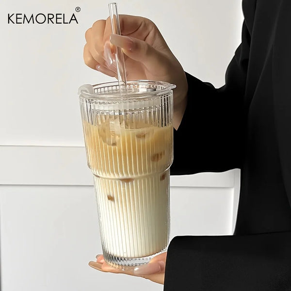A hand is pouring a light brown iced coffee drink into a glass cup with a lid.