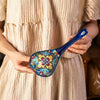 A hand holds a blue ceramic serving spoon with floral patterns against a light beige dress.