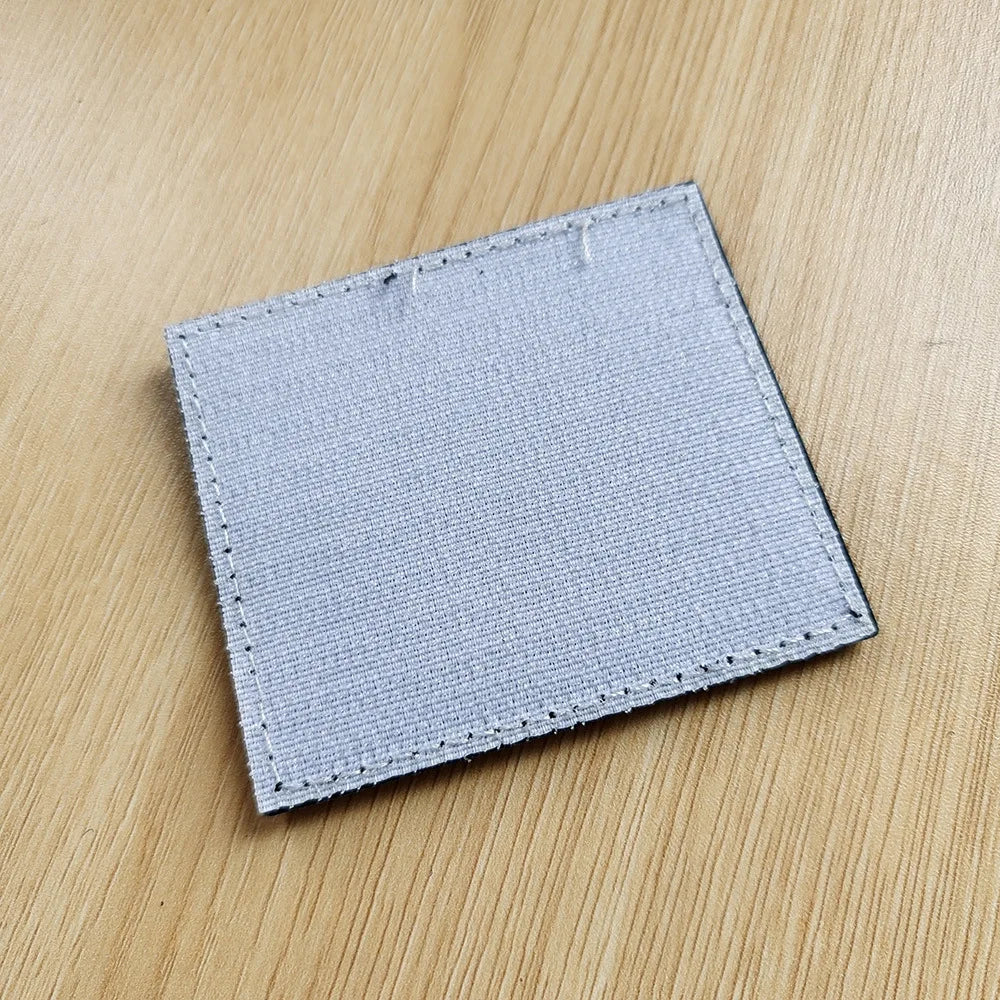 A square gray tactical PVC warning patch with a stitched border lying on a wooden surface.