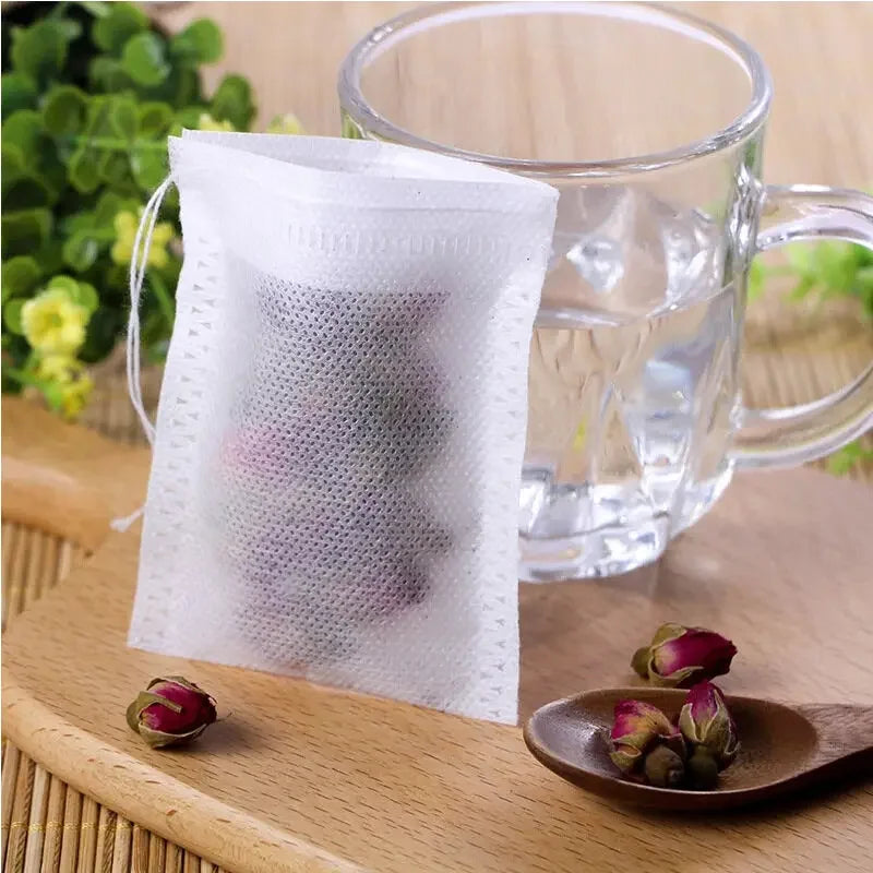 A white disposable non-woven tea filter bag is placed next to a glass mug filled with clear liquid on a wooden surface.