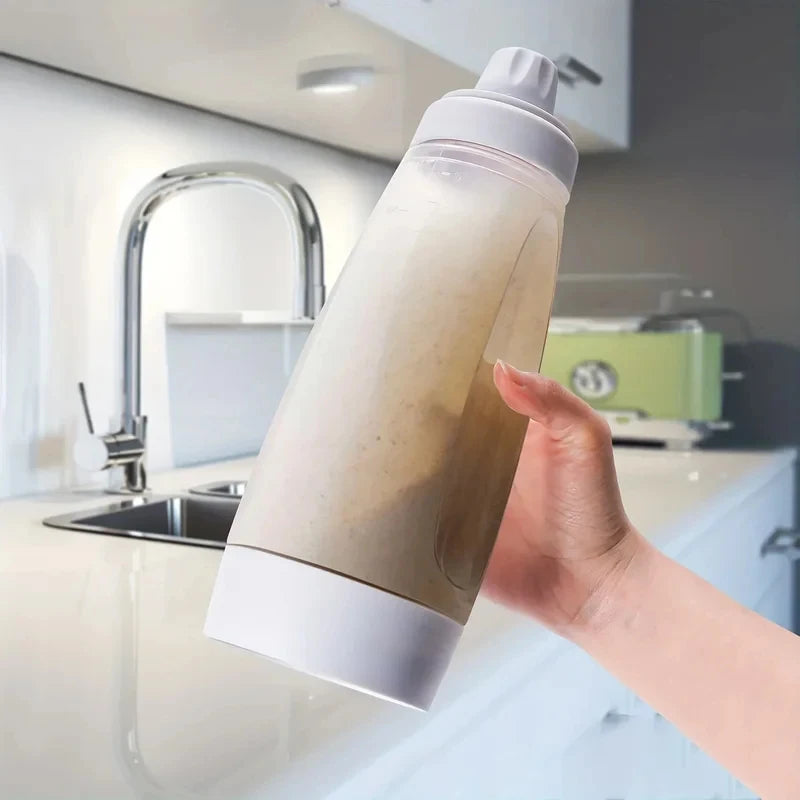 A hand is holding a clear plastic pouring bottle filled with light brown liquid, likely a baking ingredient, in a kitchen set