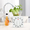 A white square mechanical kitchen timer is shown next to a glass pitcher and a faucet in a kitchen setting.