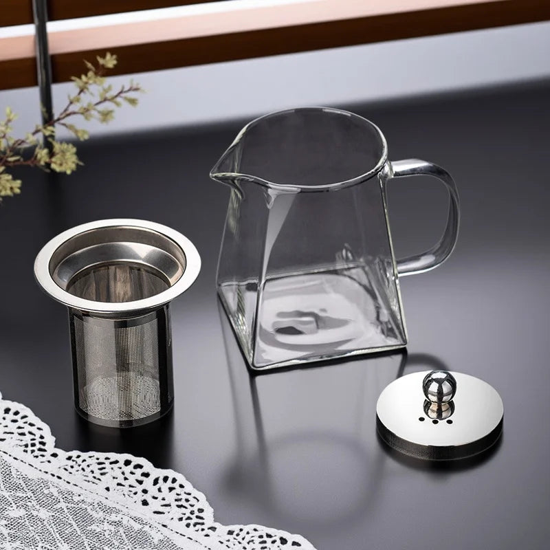 A clear glass teapot with a handle and a stainless steel infuser resting on a dark surface next to a lace doily.