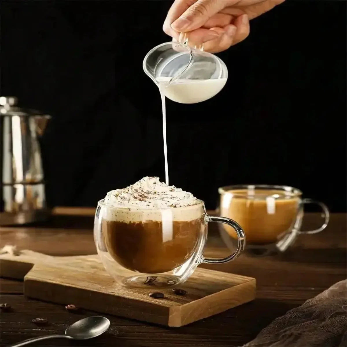 Una mano vierte leche en una taza de café de doble pared, colocada sobre una tabla de madera.