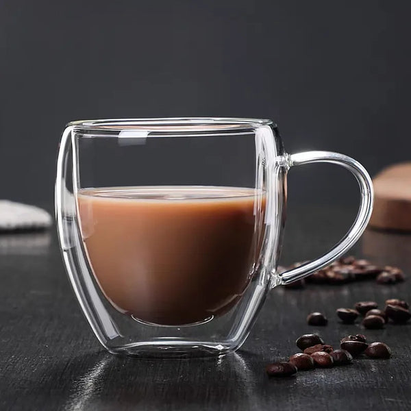 Klarer doppelwandiger Borosilikatglas-Kaffeebecher mit brauner Flüssigkeit, auf einer dunklen Oberfläche mit Kaffeebohnen.