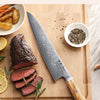 A set of seven Damascus steel Japanese kitchen knives displayed on a wooden cutting board with meat and spices.