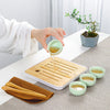A hand pours tea from a ceramic teapot onto a wooden tray and small cups for a tea ritual.