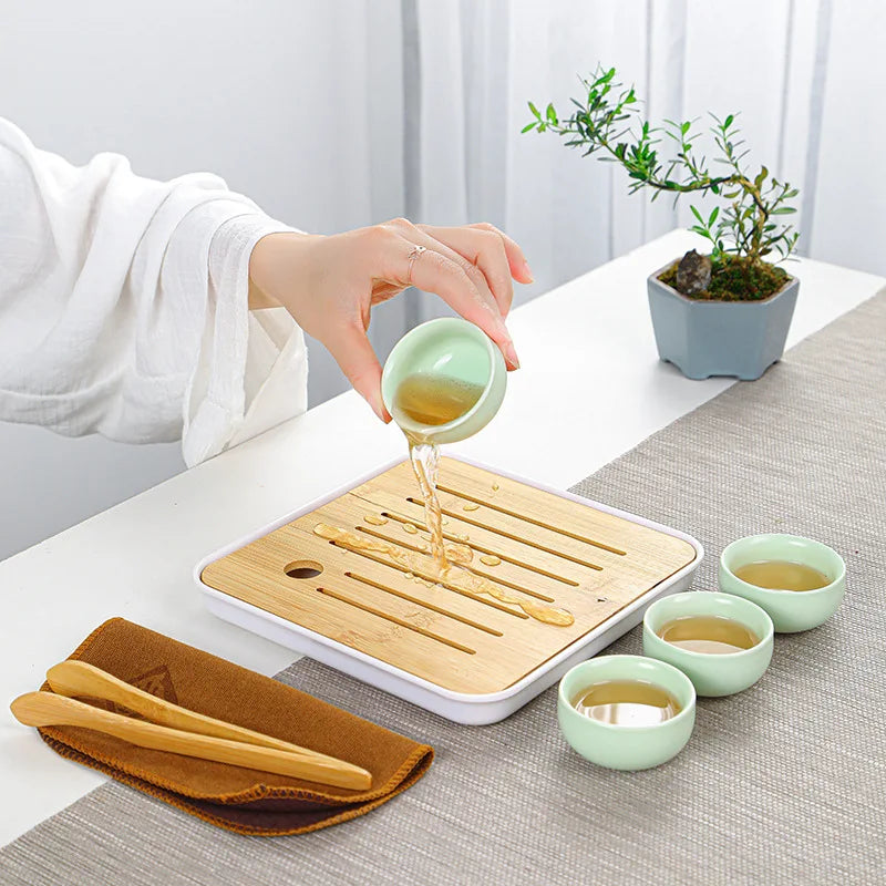 A hand pours tea from a ceramic teapot onto a wooden tray and small cups for a tea ritual.