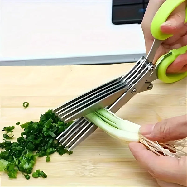 A hand is using a triple-blade stainless steel herb scissors to chop green herbs on a wooden cutting board.