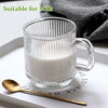 A clear glass mug with vertical stripes filled with milk, placed on a white tray next to a gold spoon.