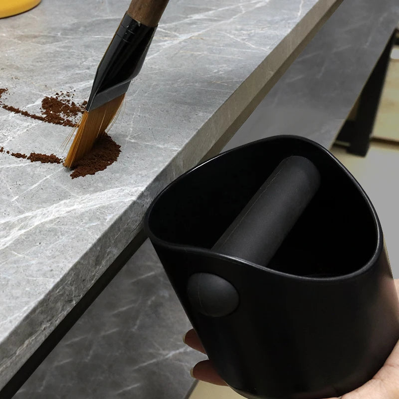 A black container holds coffee grounds, positioned next to a marble countertop and a wooden brush.