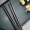 A set of five black gold alloy chopsticks resting on a dark slate board with Japanese-style sushi visible.