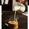 A hand pours milk from a glass pitcher into a mug on a wooden table.